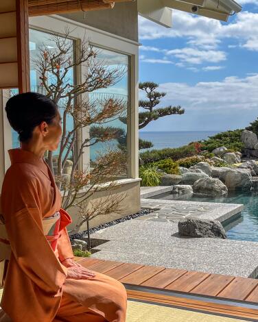 A woman in a kimono kneels outsidea house, the Pacific Ocean visible in the distance