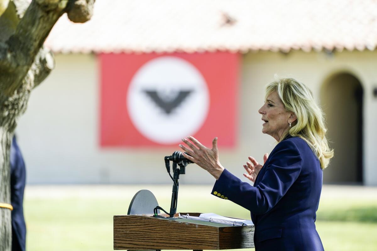 A woman gestures as she speaks outdoors at a lectern on a sunny day.