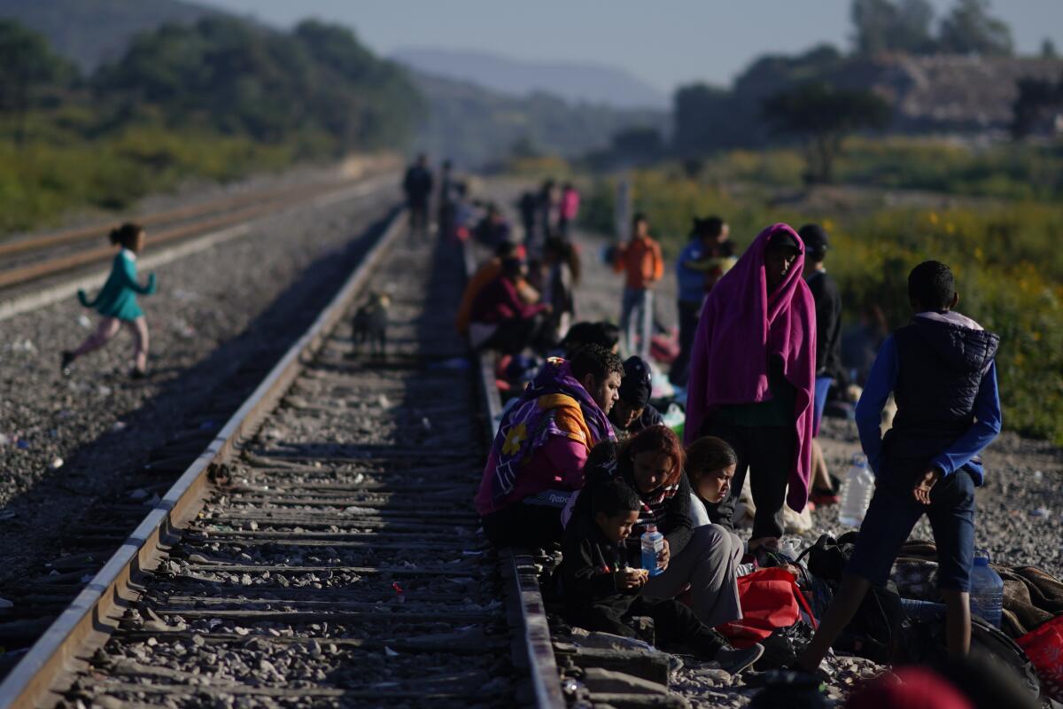 People wait with children and belongings next to railroad tracks.