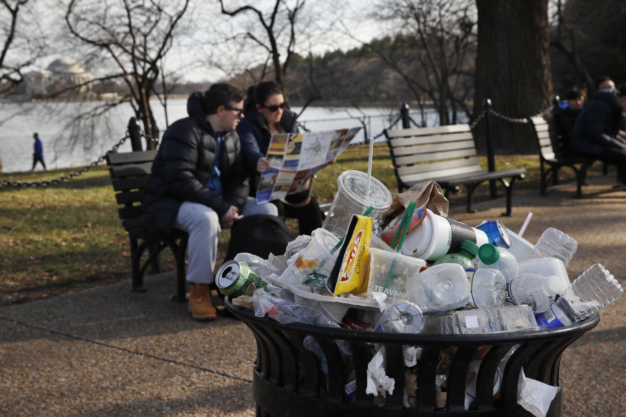 A couple sits on a park bench near an overflowing trash bin.