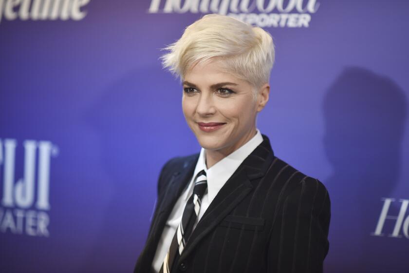 A woman with short platinum blonde hair in a dark suit and a striped tie standing in front of a blue backdrop