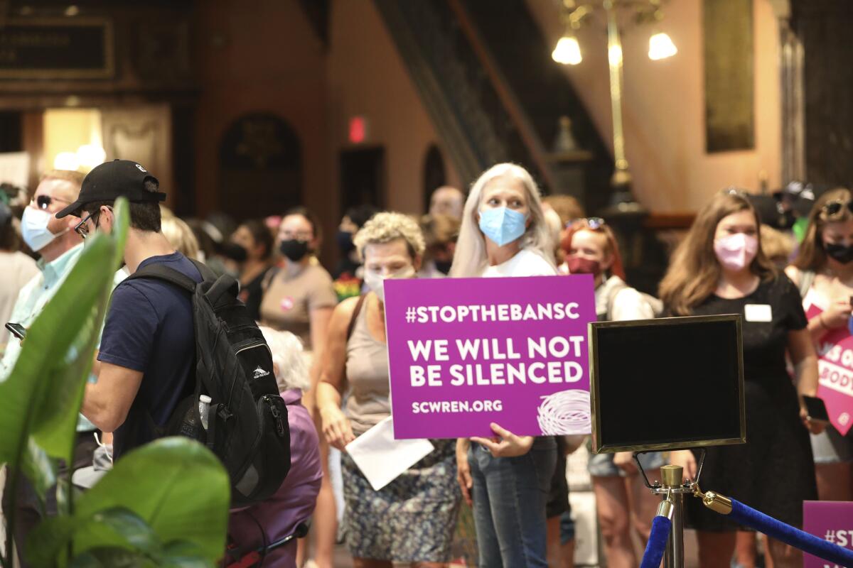 A crowd of abortion rights supporters in a building lobby, one with a sign reading "#StopTheBanSC / We will not be silenced."