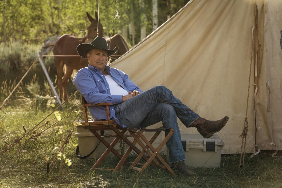 A man wearing denim and a cowboy hat sits in a director's chair in front of a tent and horses