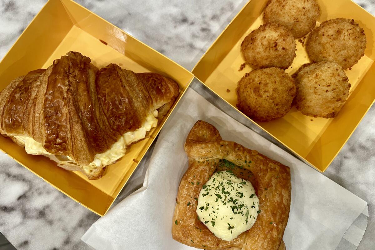Croissant egg salad sandwich and chicken bites in yellow to-go boxes and a special croissant on a flattened paper bag