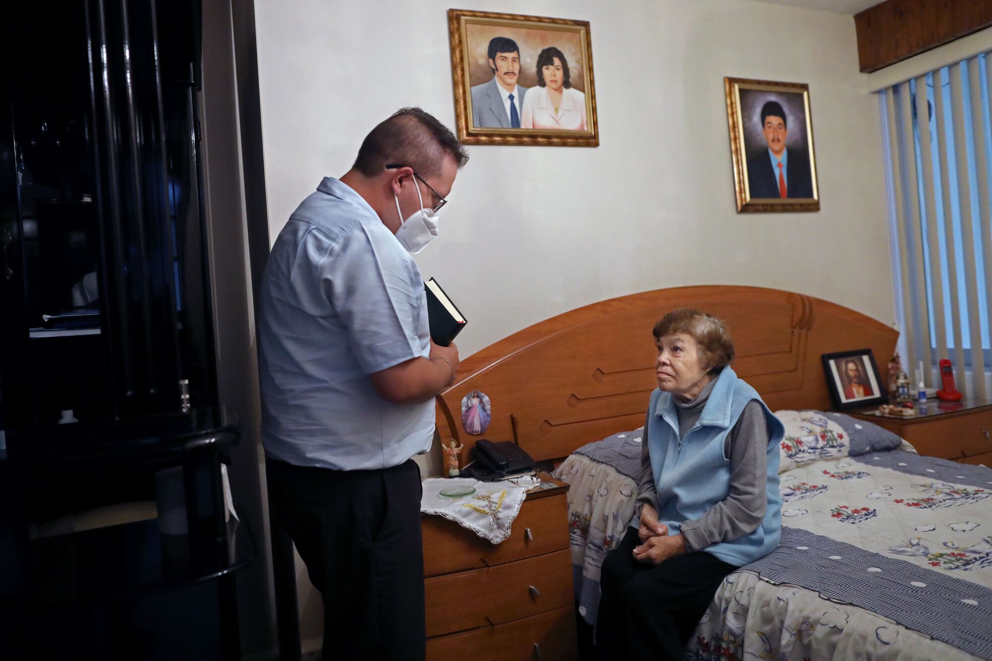 A woman sits on her bed, talking to a priest standing near her in a mask