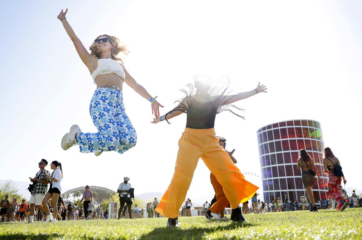 Woman in white top and blue flower pants jumping next to woman in black top and yellow pants in grass at Coachella
