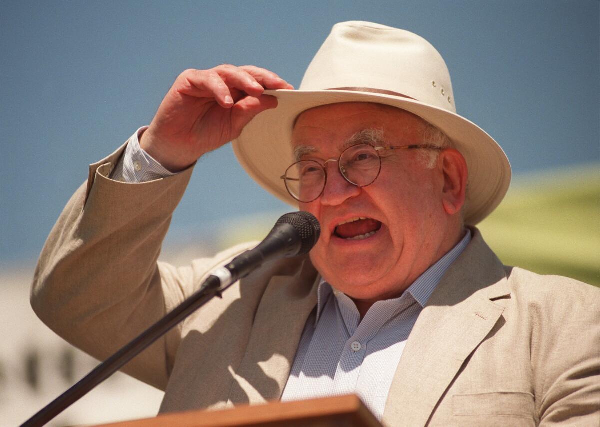 Ed Asner in a wide-brimmed hat delivering a speech at a podium
