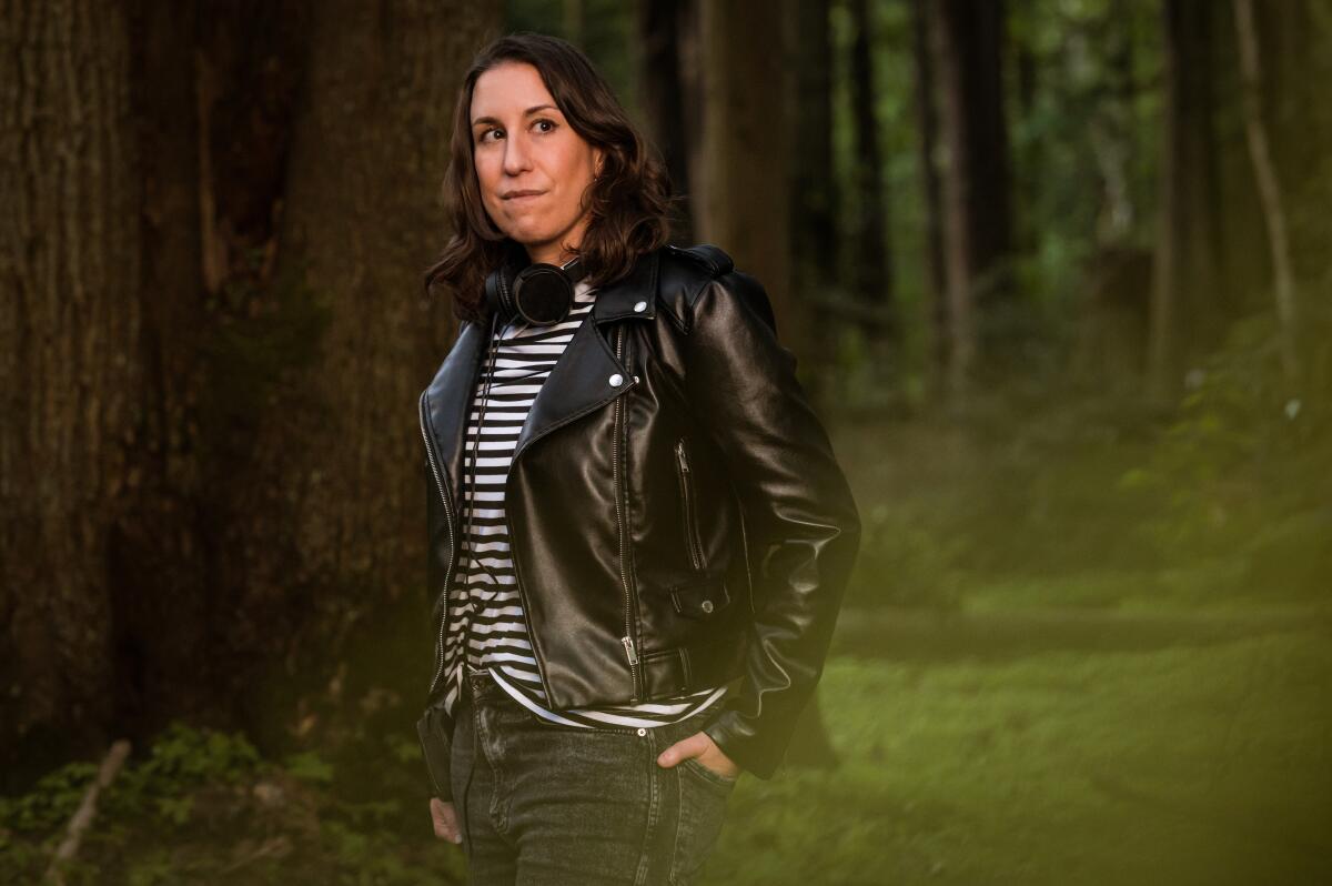 A woman wearing a striped shirt and black jacket stands in the woods.
