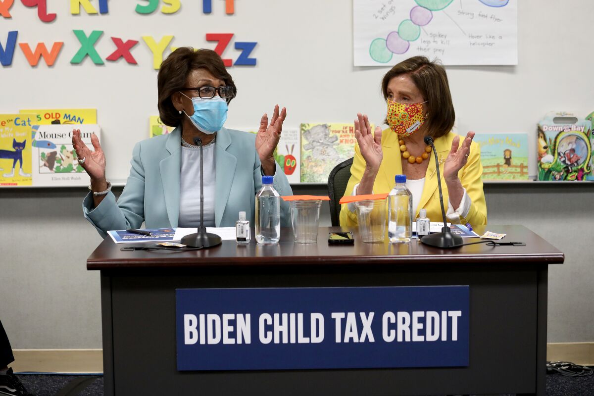 Maxine Waters and Nancy Pelosi sit at a desk with a whiteboard and children's books behind them. 