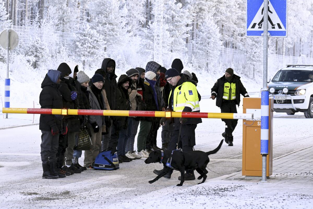 Migrants stand in the snow as a customs official tends to a dog on a leash 