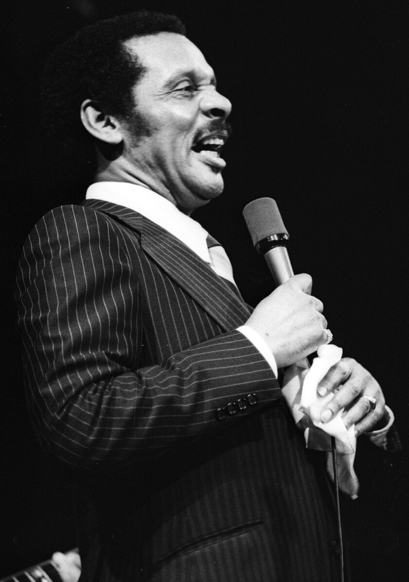 A man in a pinstripe suit stands, smiling and holding a microphone