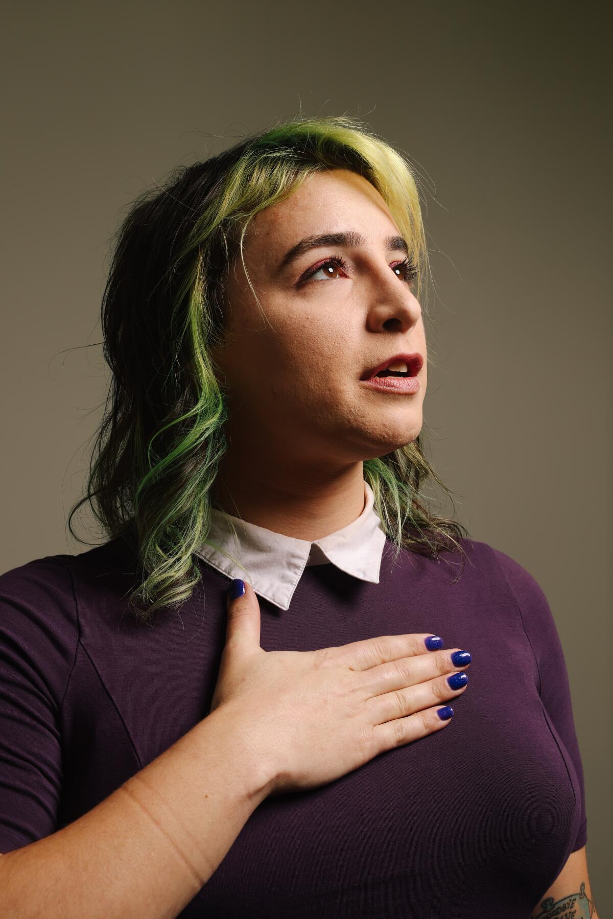 A woman with green hair and a purple shirt poses with her right hand on her chest 
