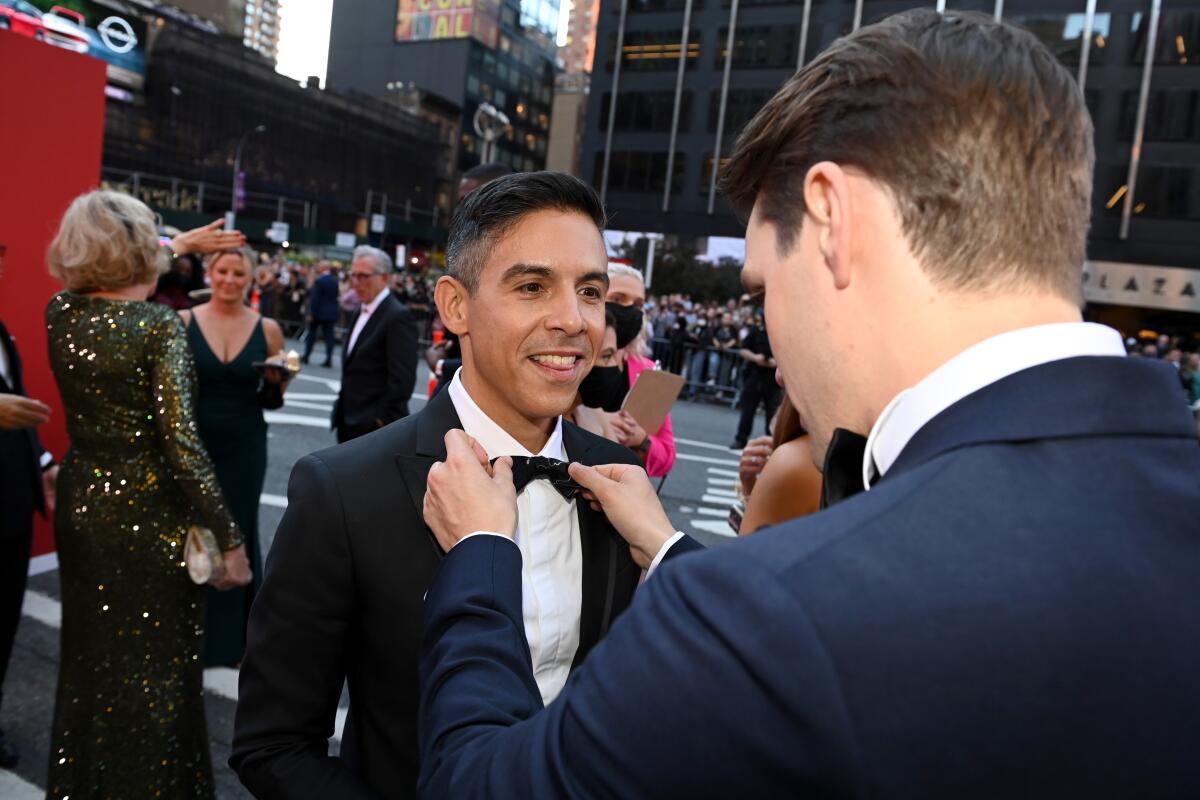 A man in a tuxedo smiles as another man straightens his bow tie while standing on a street among other dressed-up people