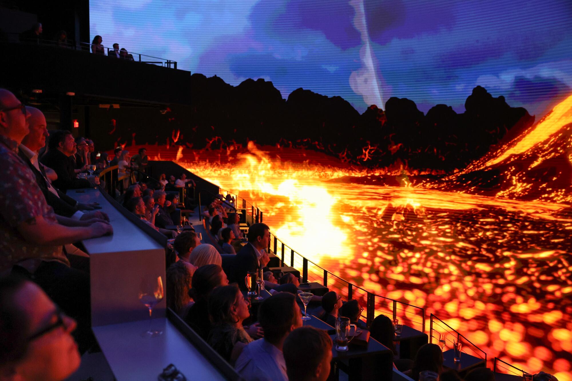 Seated audience members watch what appears to be bubbling lava under a cloudy sky