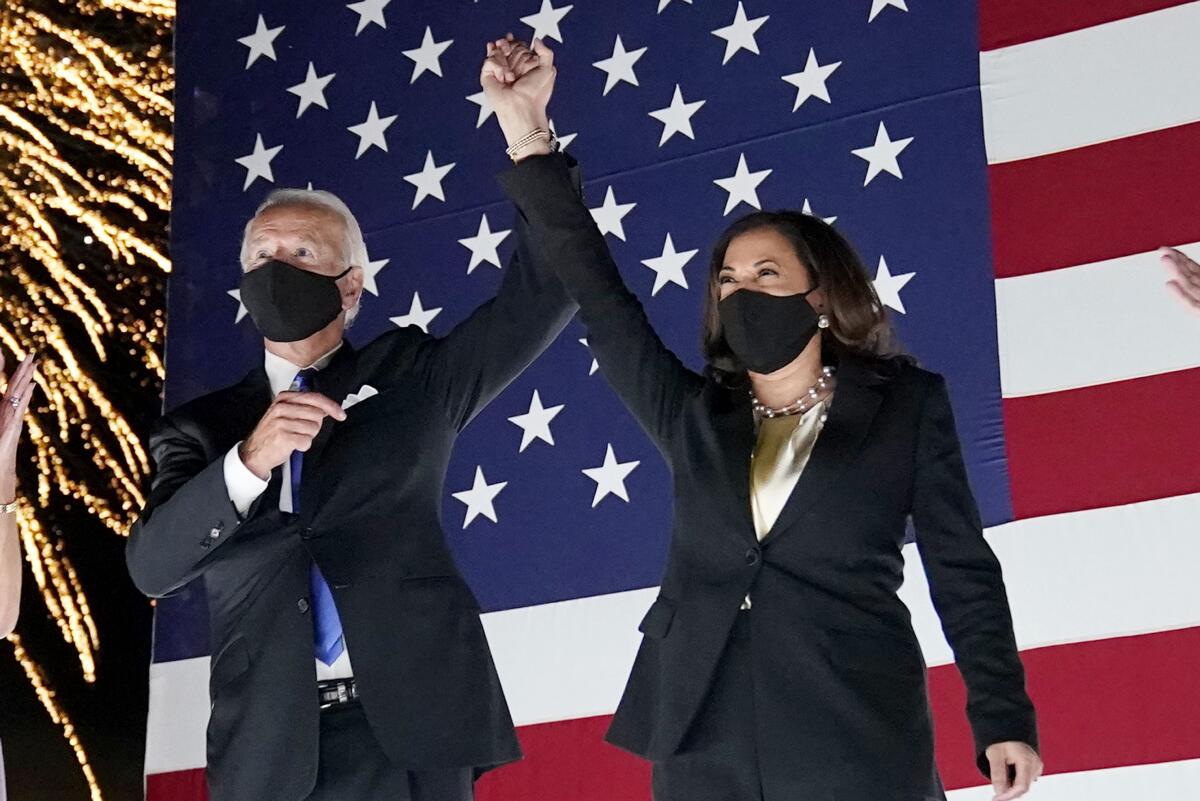 Joe Biden and Kamala Harris raising their joined hands while wearing face masks in front of an American flag.