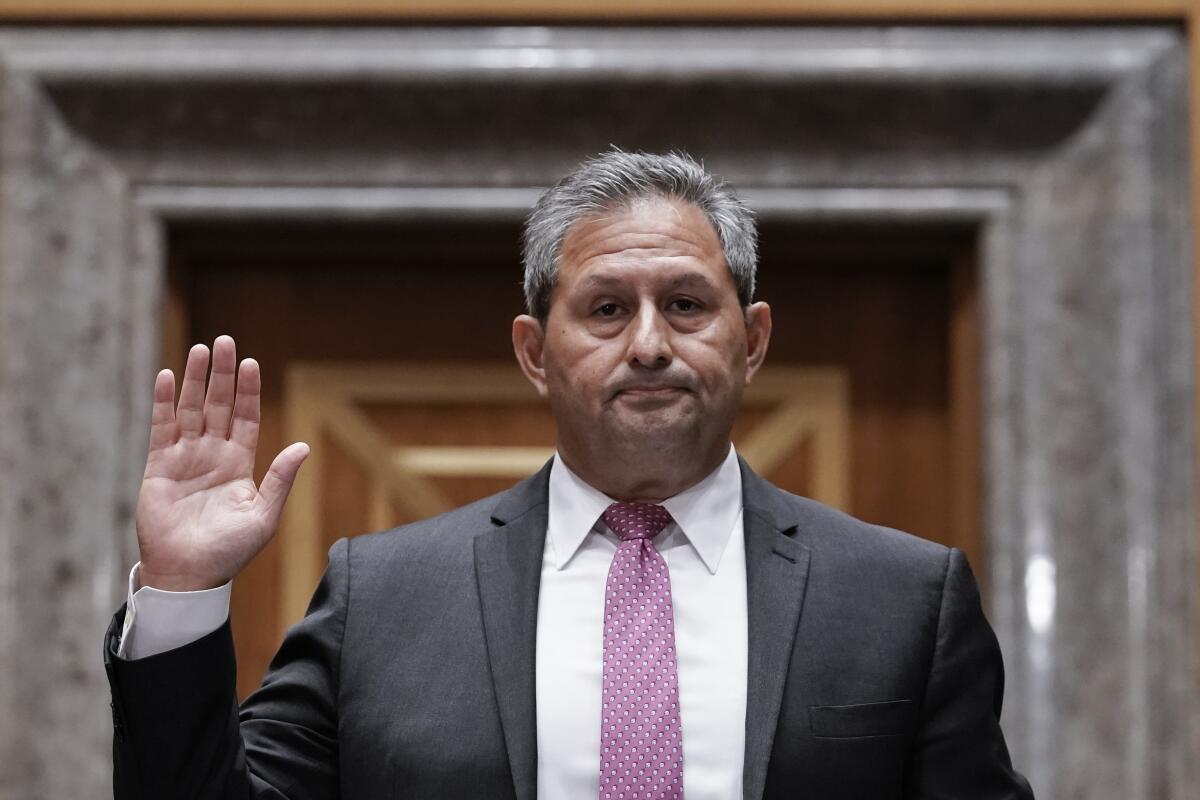 A man in suit and tie has a grim expression on his face as he holds up one hand. 