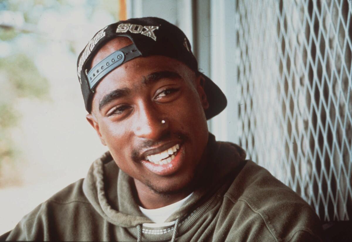 Tupac Shakur, wearing a hoodie and a backwards baseball cap, smiling.
