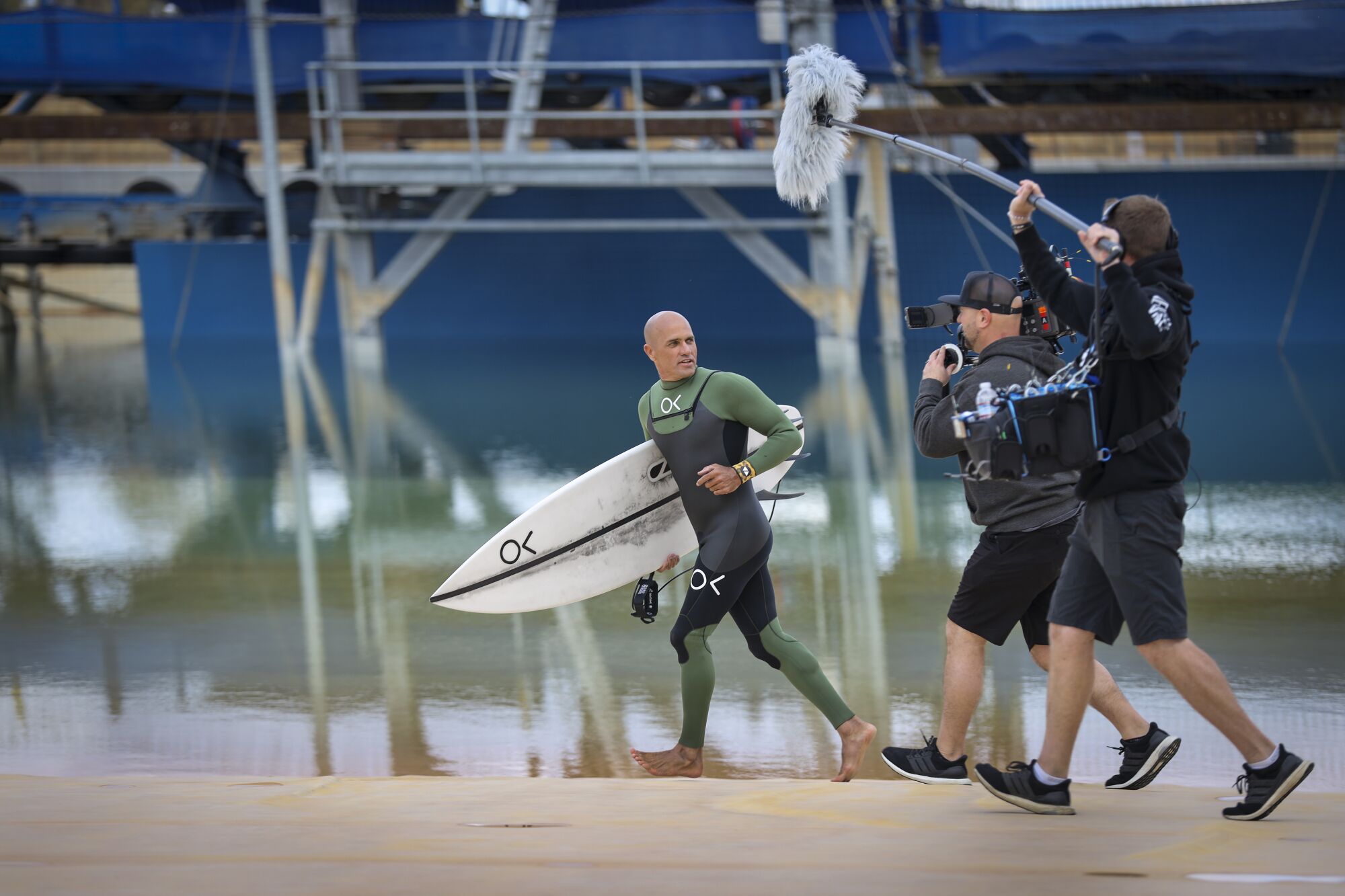 Kelly Slater runs with surfboard in hand as a camera crew follows