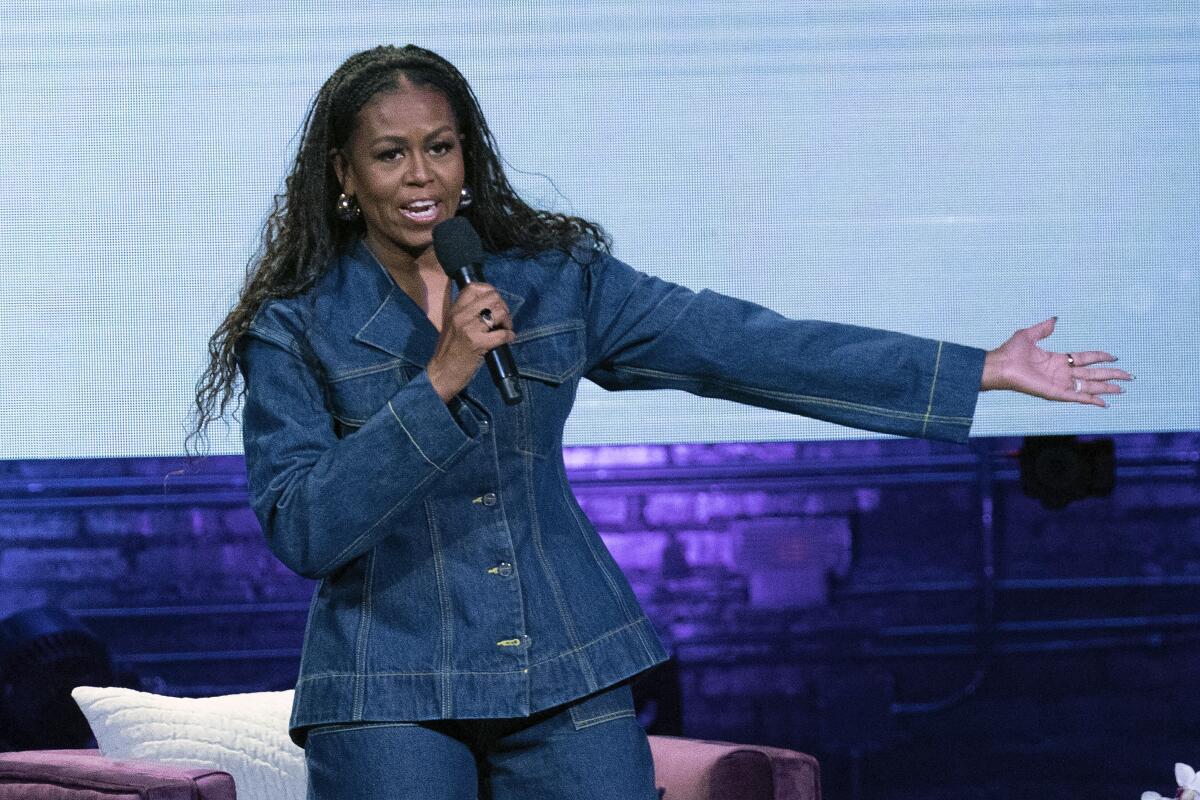 Former First Lady Michelle Obama speaking on a stage holding a microphone in a denim outfit