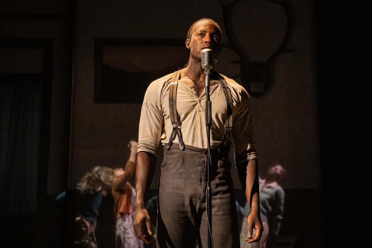 An actor in simple old-fashioned shirt, pants and suspenders stands at a microphone onstage
