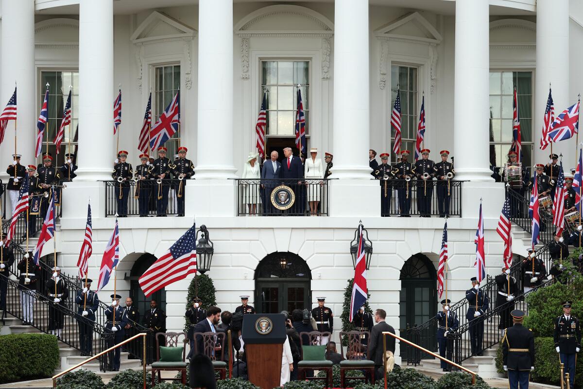 Two men in suits and ties, flanked by two women wearing white, stand on a white balcony, flanked by people in dark uniforms