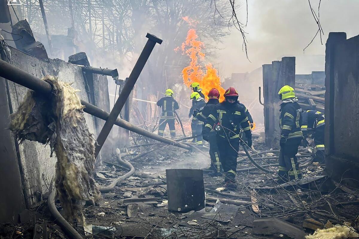 Emergency workers walk amid rubble as flames rise behind them. 