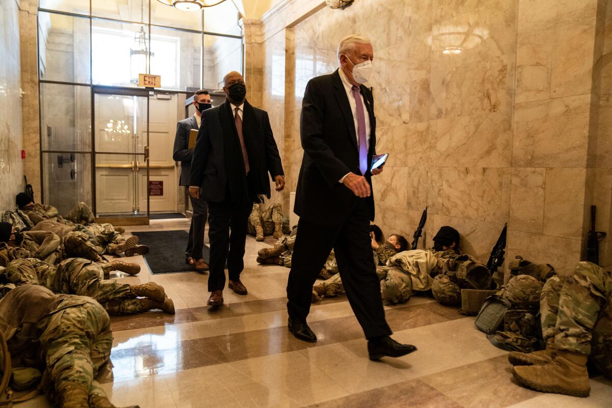 Three men in suits and masks walk along a hallway lined by sleeping men in camouflage.