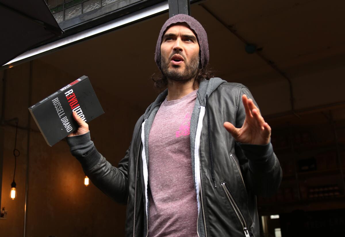Russell Brand in a purple T-shirt, gray hoodie and a beanie holding up both hands with a book in his right hand