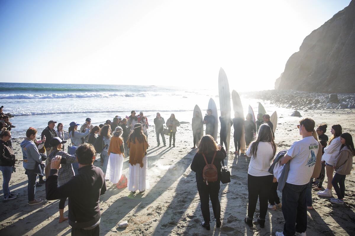People gather in a circle on a sunny beach, including seven surfers holding their boards.