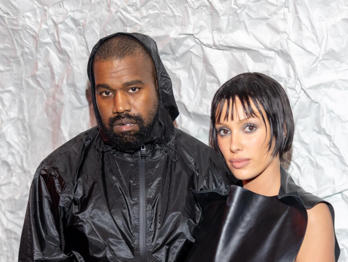 Kanye "Ye" West in a black hoodie standing next to a woman with short hair and bangs in a black sleeveless shirt