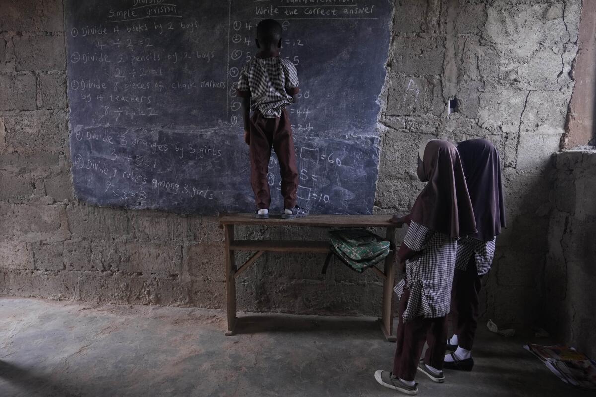 A child stands on a table to write on a blackboard in a spartan, dimly lit classroom