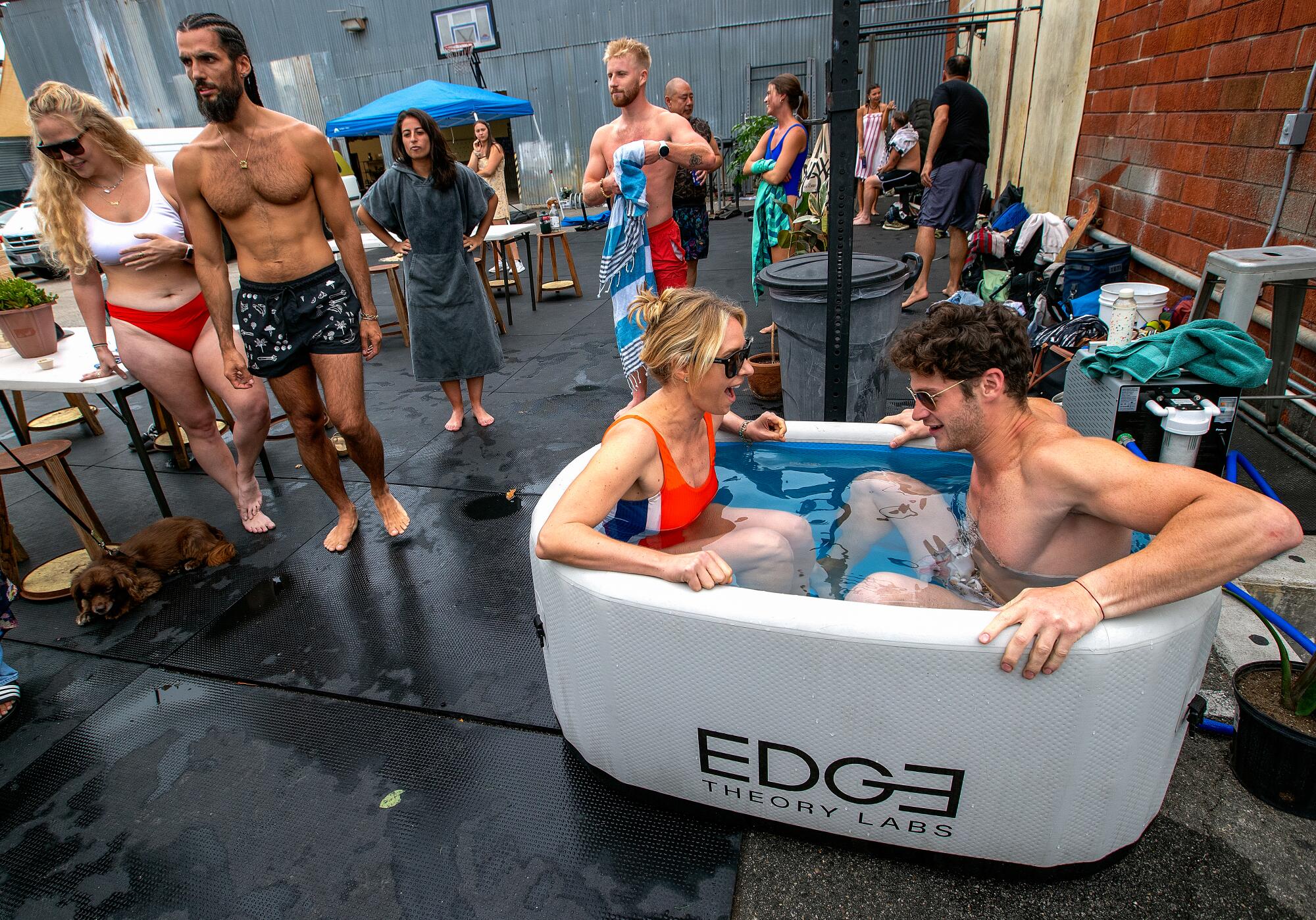 Two people sit in an outdoor ice bath while others in bathing suits stand nearby.