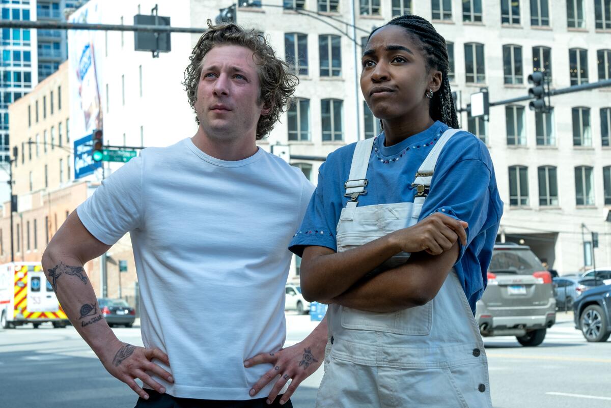 A man in a white T-shirt stands next to a woman in a blue shirt and overalls.