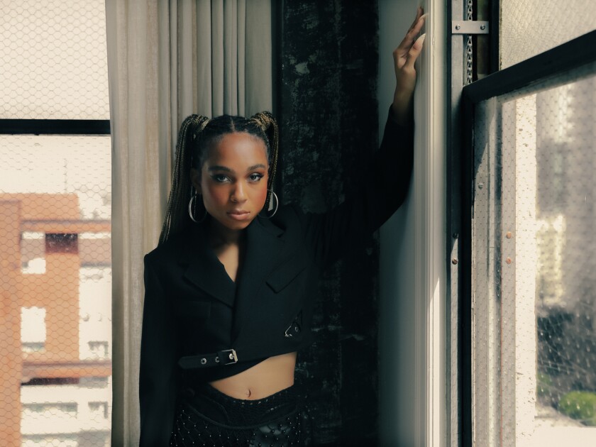 Eva Reign stands against a window, one hand up on the adjacent wall.