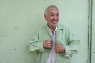A man with grey hair wearing a mint green jacket smiles before a mint green wall