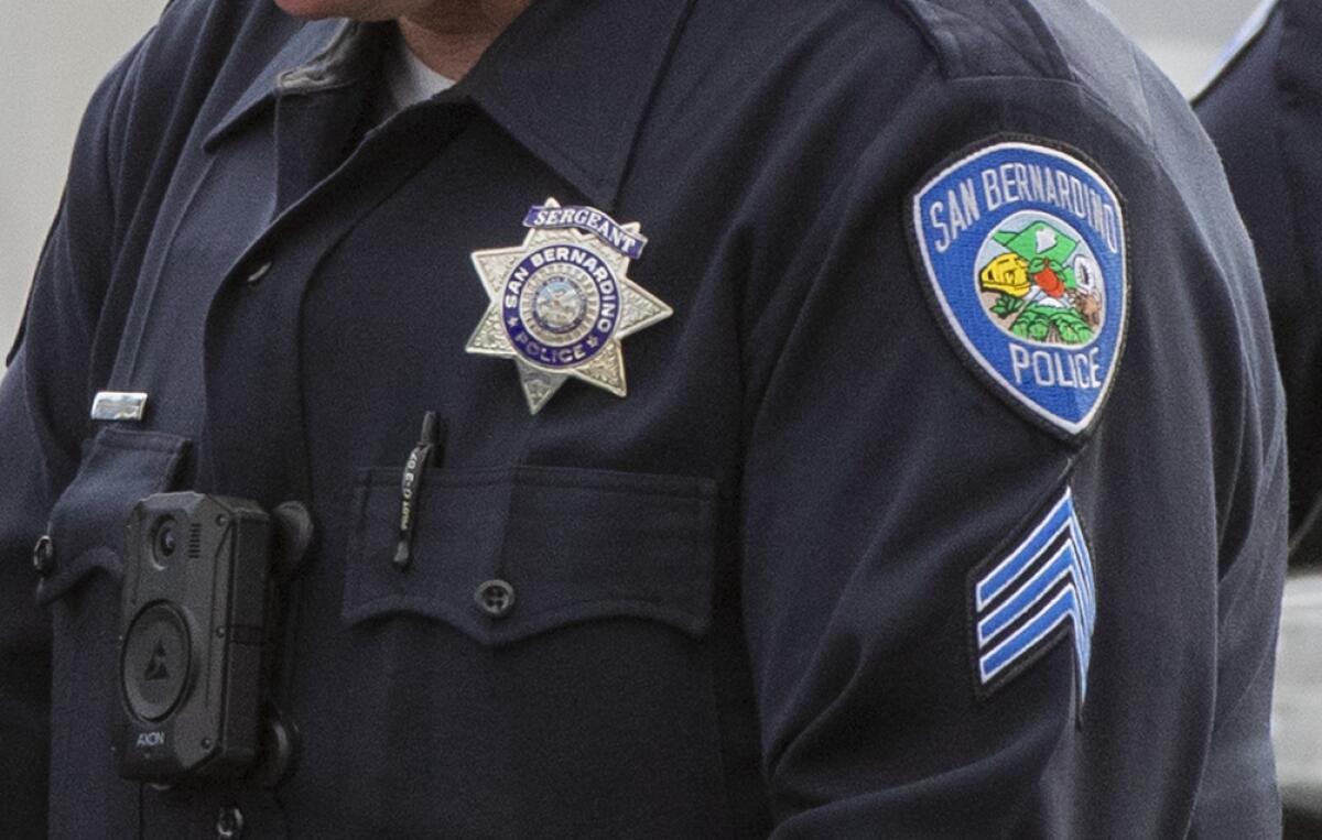 The chest of a person in a navy uniform with a silver badge and sleeve patches, one reading "San Bernardino Police"