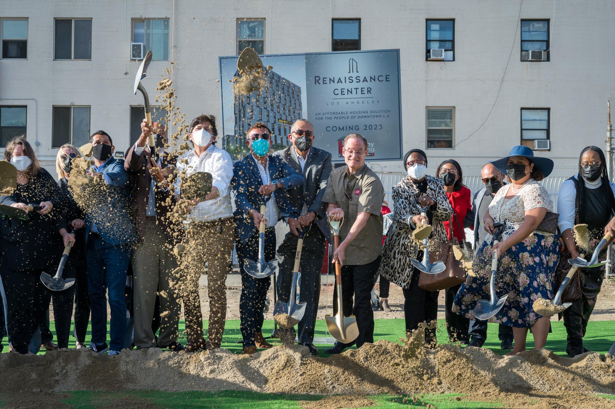 A line of people dig up and throw dirt in the air during an outdoor groundbreaking ceremony.