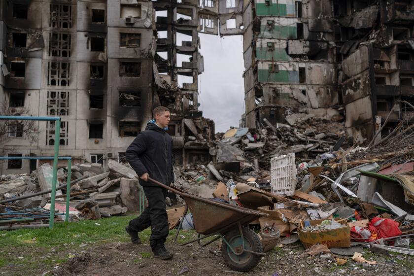 A young man pushes a wheelbarrow in front of a destroyed apartment building