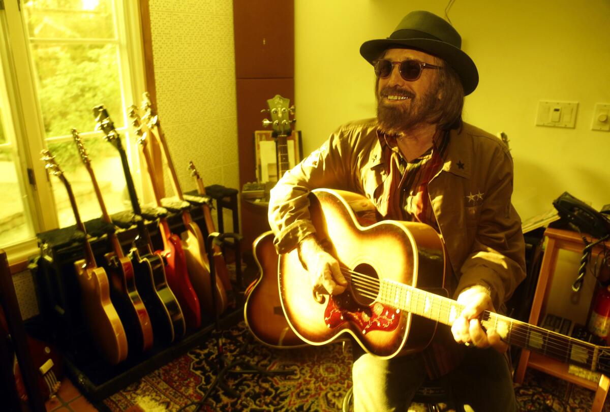 Tom Petty, dressed in a jacket, sunglasses and fedora, smiles and plays a guitar while seated in his home.