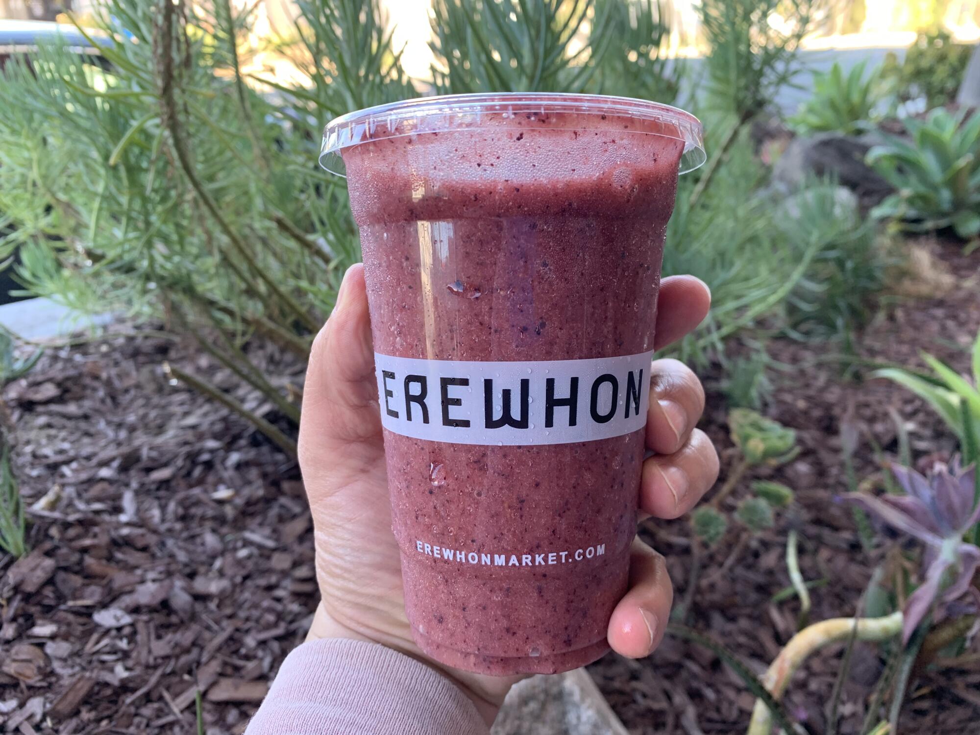 A hand holds a red smoothie in a plastic cup that says Erewhon.