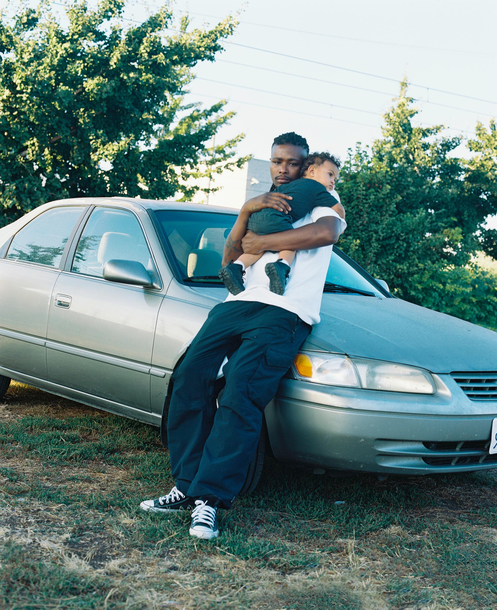 A man leans against a car, holding a baby in his arms.