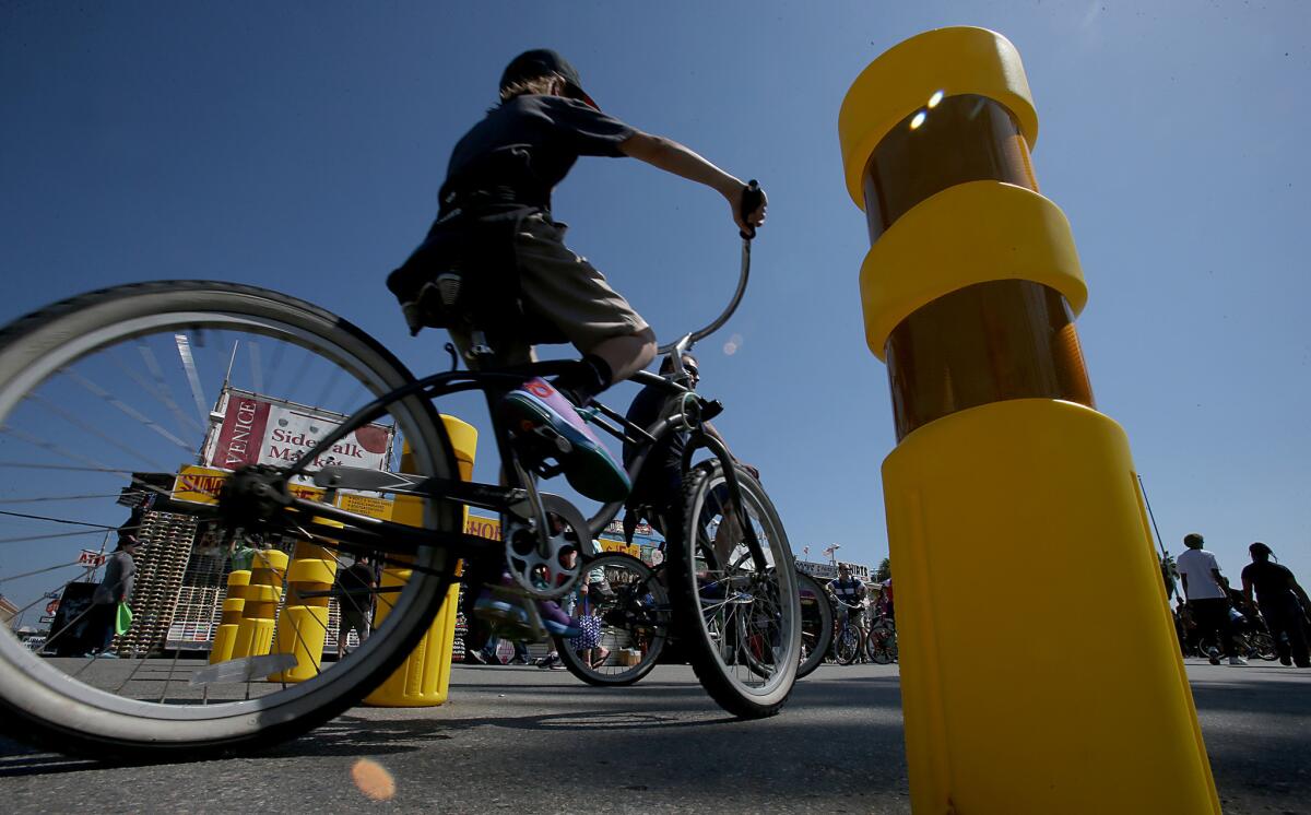 Cyclists and strollers on Venice Beach pass bright yellow bollards that have been installed along the boardwalk.