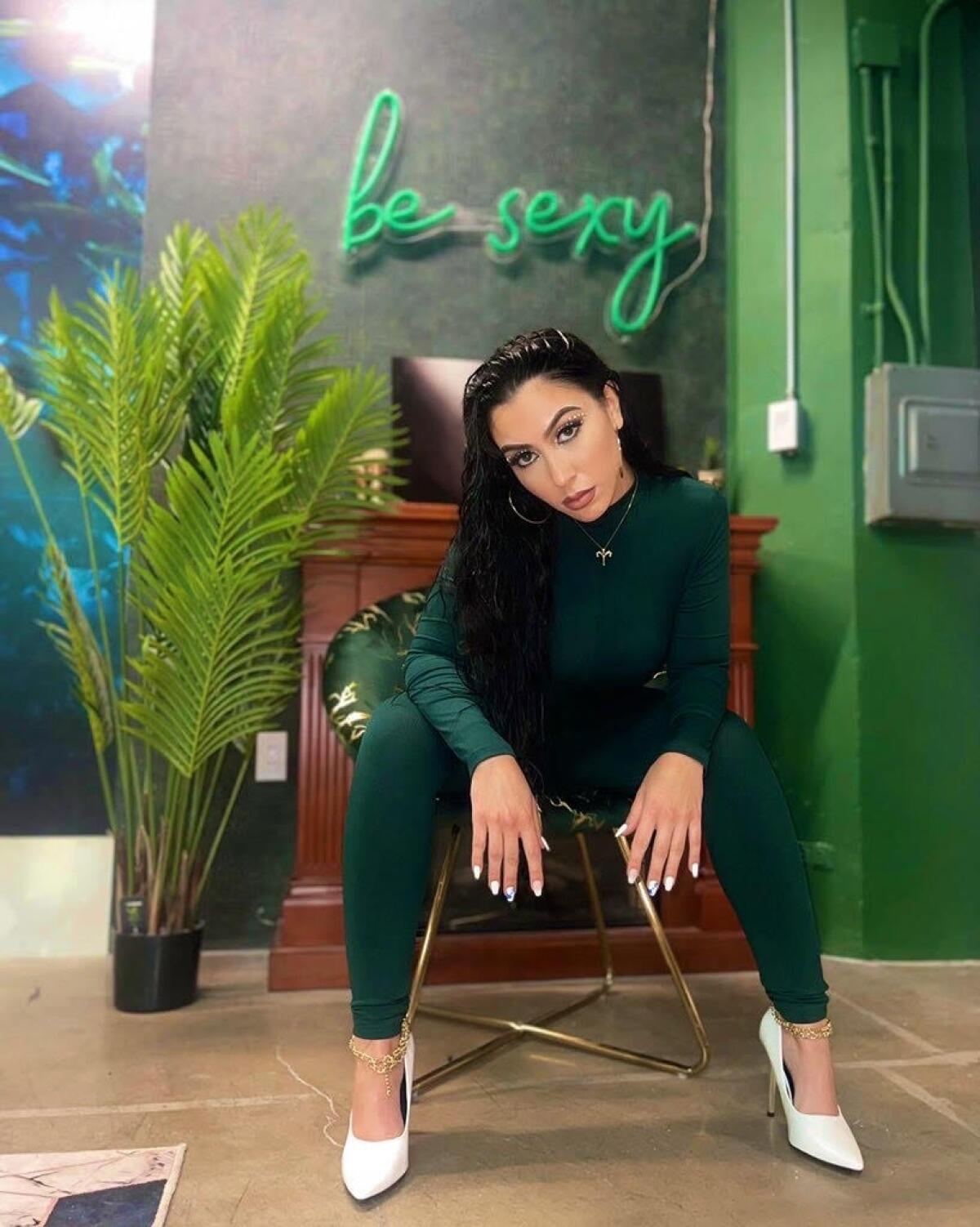Jessica Vanessa, a social media influencer, sits in a chair beneath a neon sign reading "be sexy."