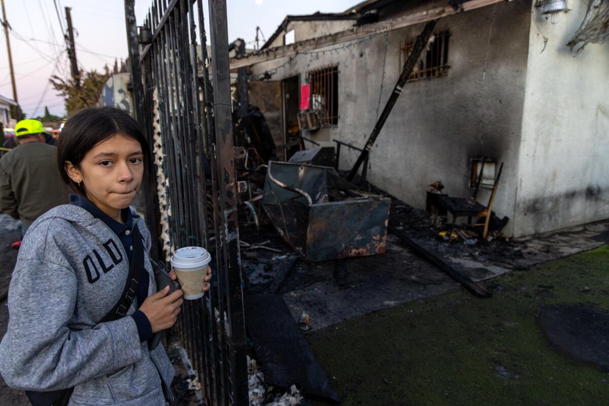 A girl holding a coffee cup and standing outside a fence next to a burned-out home and charred debris