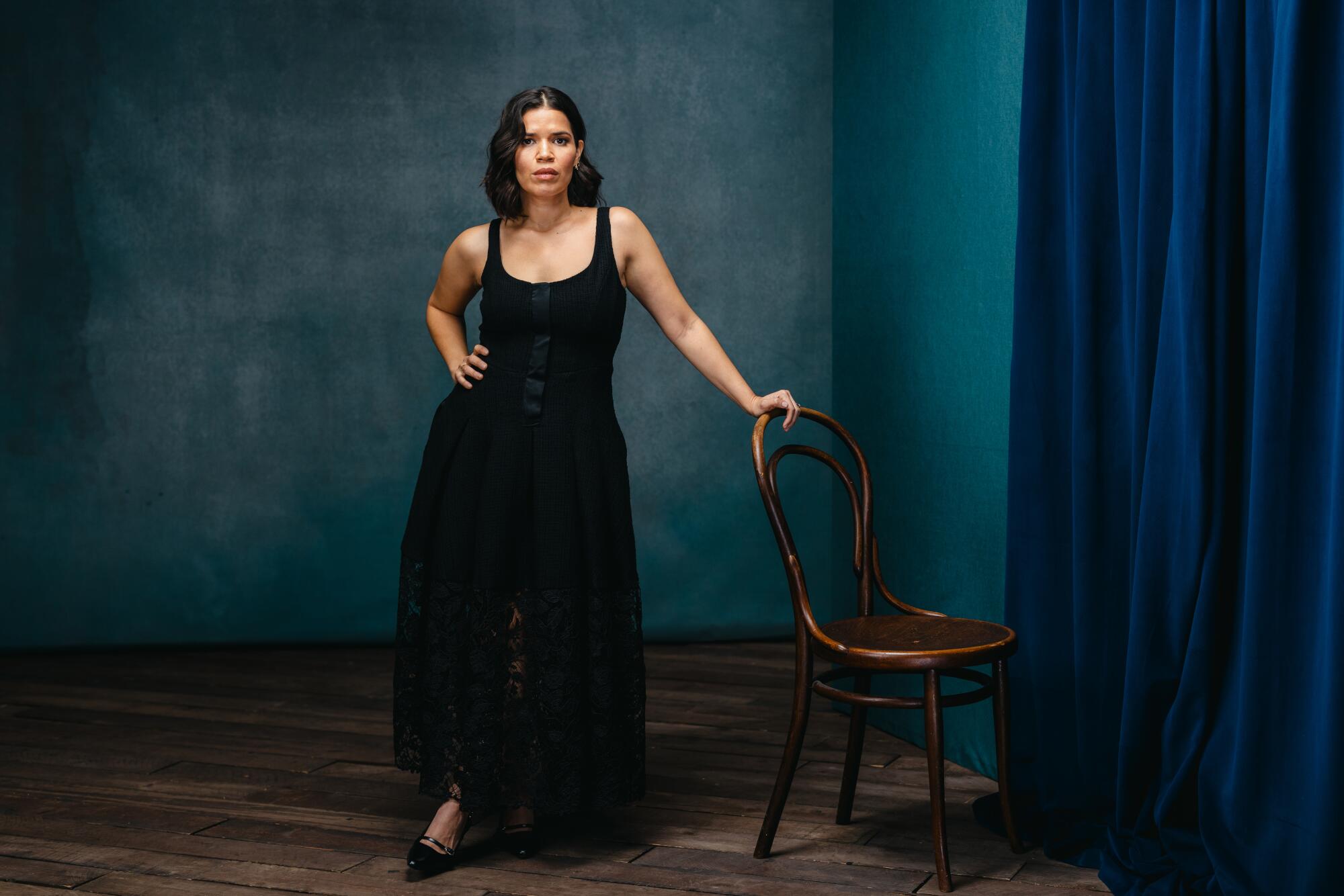 America Ferrera leans on a wooden chair, on hand on her hip for a portrait.