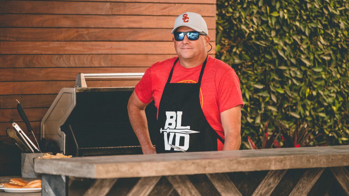 USC coach Clay Helton wears an apron that says "BLVD" as he stands at an outdoor grill.