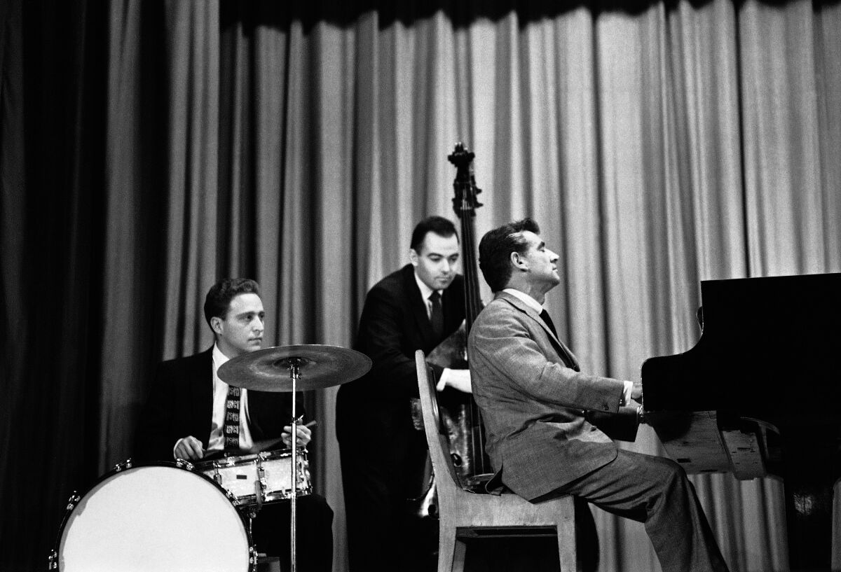 Black and white photo of a man at right playing a piano with a man, far left, playing drums, and another, middle, bass