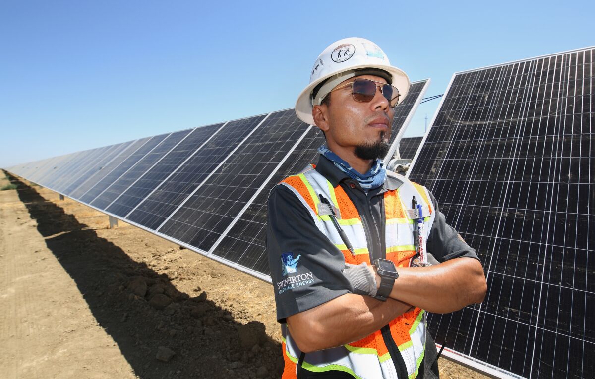 A construction worker in white hat and a vest stands, arms crossed, next to a row of dark solar panels 