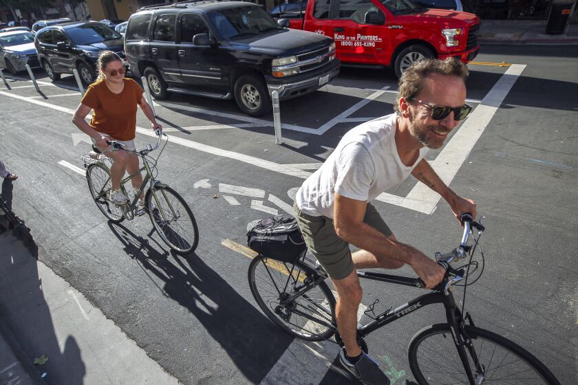 Dan Blanchett and his wife Goda Blanchett ride bicycles on a bike lane next to where cars park away from the curb, upper left, on J Street, at the Eleventh Avenue intersection in downtown on Tuesday, July 9, 2019 in San Diego, California.