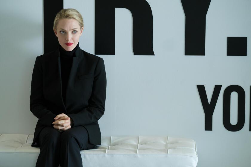 A woman in a black suit with red lipstick sits on a white bench against a white wall with black letters on it.